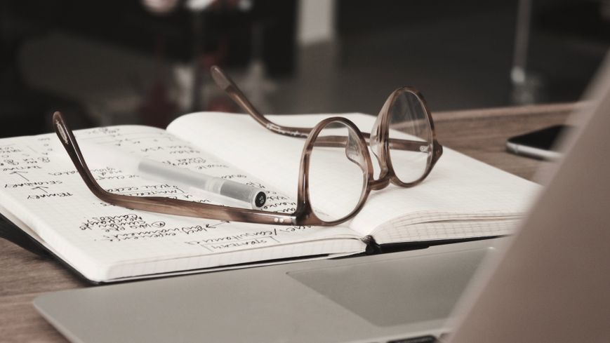 Kahverengi masa üstünde araştırma notları içeren bir defter ve üzerine koyulmuş kalem ile gözlük