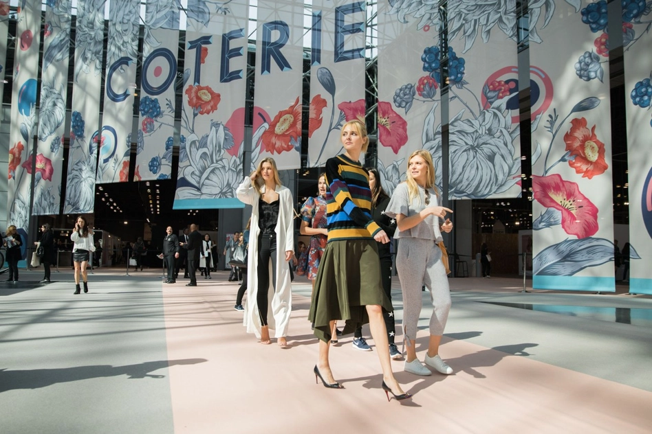 Three women walking in front of the Coterie banners in a fashion trade show.
