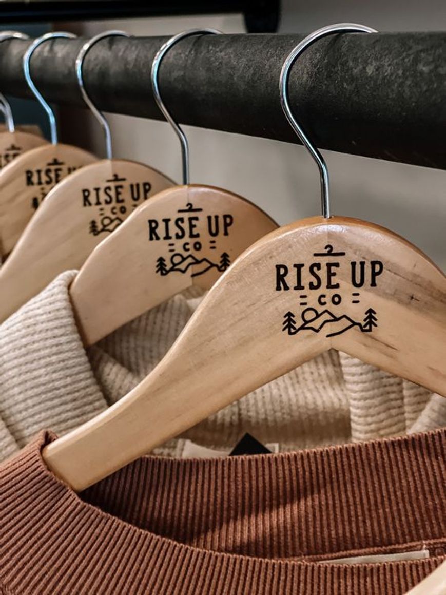 Cinque grucce in legno con la scritta «Rise Up» all'interno di una piccola boutique di abbigliamento